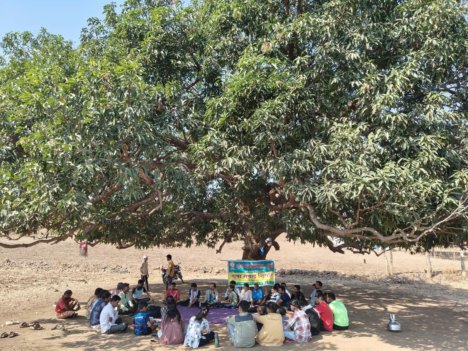 यंग फाऊंडेशनच्या कार्यक्रमासाठी मोठ्या झाडाच्या सावलीत बसलेले लोक.