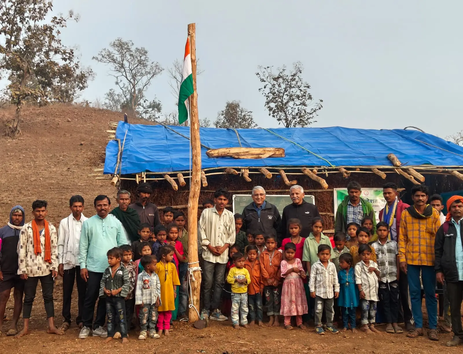 सातपुडा टेकड्यांमध्ये एका झोपडीसमोर झेंडावंदन करताना लोक आणि मुले उभे आहेत.
