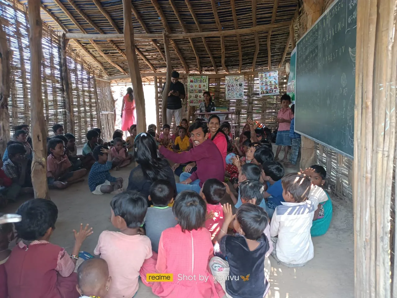 यंग फाऊंडेशनच्या बांबू आणि लाकडाच्या वर्गात मुले आणि शिक्षक एकत्र बसले आहेत.