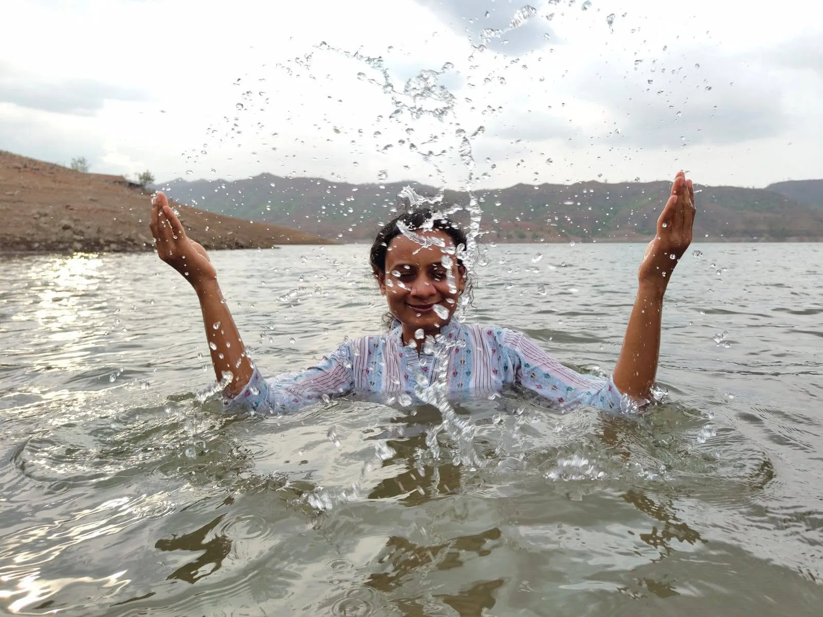 एक महिला पाण्यात उभी आहे आणि तिने आनंदाने दोन्ही हात वर करून पाणी उडवले आहे.