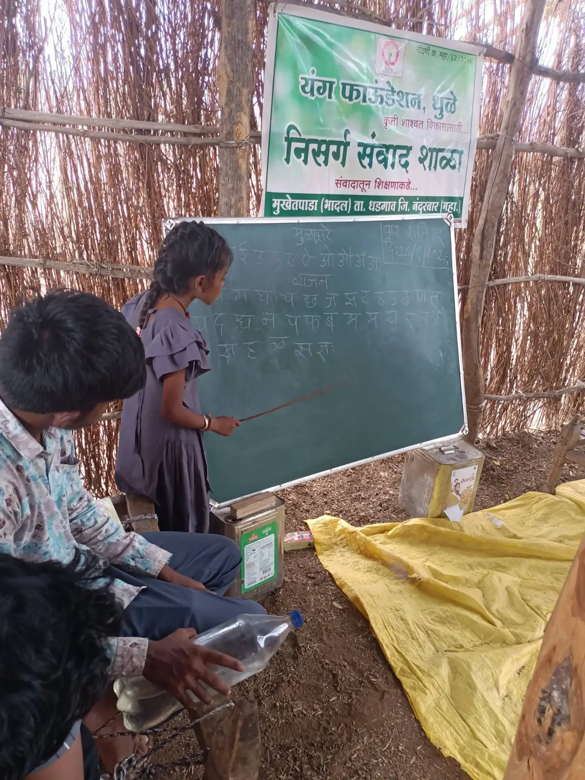 निसर्ग संवाद शाळेत फळ्याजवळ उभी असलेली मुलगी व समोर बसलेले लोक.