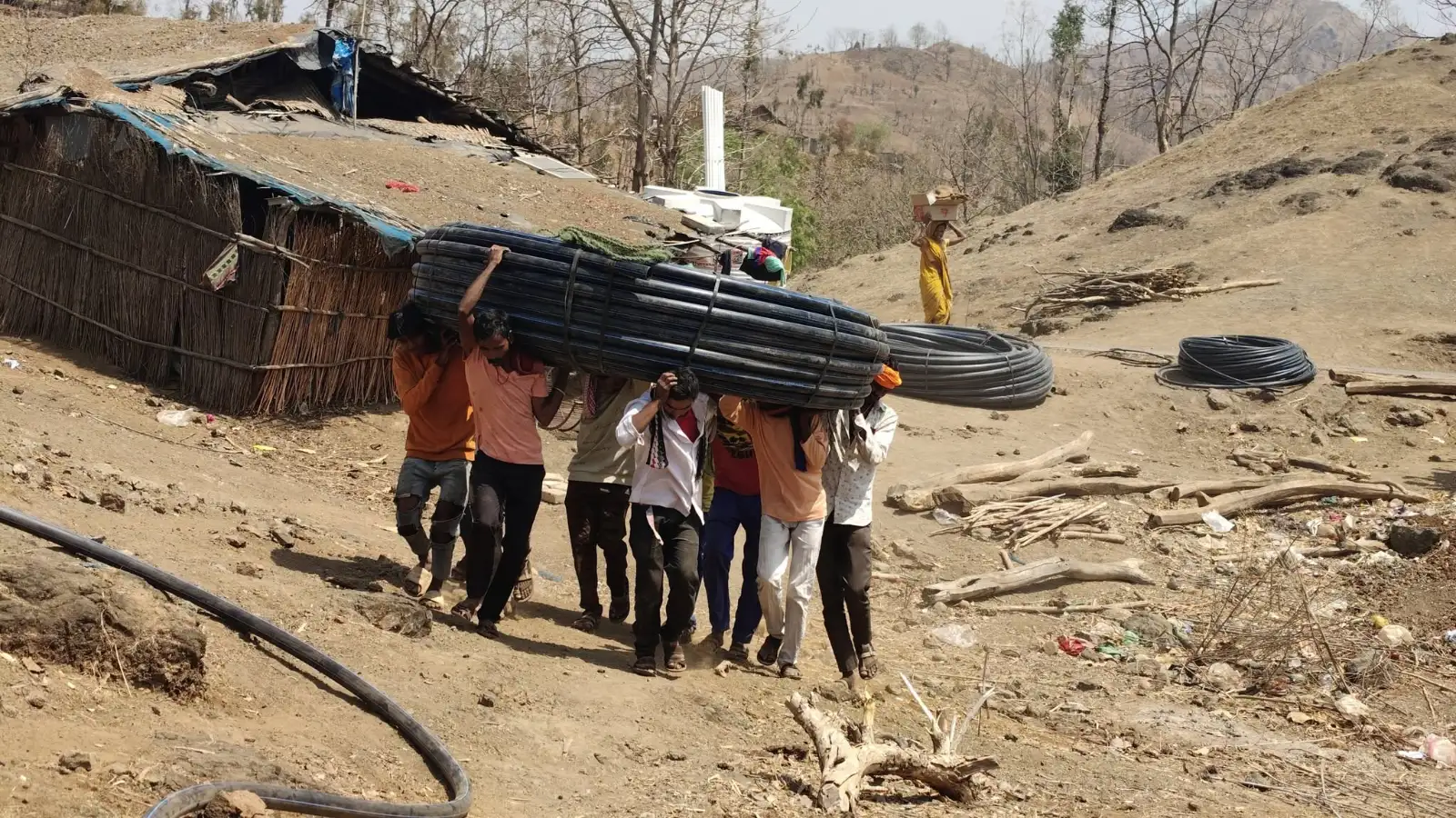 सातपुडा टेकड्यांमध्ये काळ्या प्लास्टिकच्या पाईप्सचे मोठे बंडल उचलून चालणारे लोक.